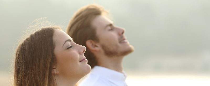 On the left in the foreground is a young women you can only see her head. She has long brown hair and has her head up looking out to the right with her eyes and mouth closed smiling like she is breathing in the fresh air. Behind her is a young man with brown hair and stubble wearing a white t-shirt doing the same. The photo is light with a pale background like the sun is shining on them.