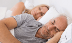 A couple sleeping in bed. closest to the front is an older man with wite hair and stubble. He's weraing a grey t-shirt with his right arm ontop of the sheets bent at the elbow. His left had is under his head. The lady asleep behind has long blond hair and you just see she has a white strappy top on, again with her left hand under her head. They are both asleep with their moths closed looking relaxed. the bedding is white as is the headboard.