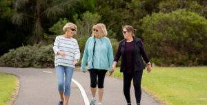 three ladies walking and chatting along a cycle/footpath. It looks like they are in a park as you can see the grass either side of the path and trees and shrubs behind them. They are all wearing sunglasses and light summer clothing. for an article about breathing for better womens health