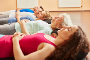 Five people are lying in a row side by side, a mixture of men and women of different ages. There heads are to the right and they are lying width ways, you can just see their upper torsos they all have their eyes closed and their hands on their upper abdomens just below the base of their ribcages. They are clearly in a studio of some kine heads on blosters and you can see the wooden floor beneath them.