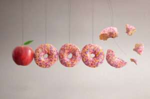 On the left is an apple that is suspended from string then to the right three pink iced donuts with 100's and 1000'2 sprinkled on top. then at the far right of them is a donut that is broken off into bits. The image gives the impression of a pendulum swing where the apple on the left has bashed the donut on the opposite side off