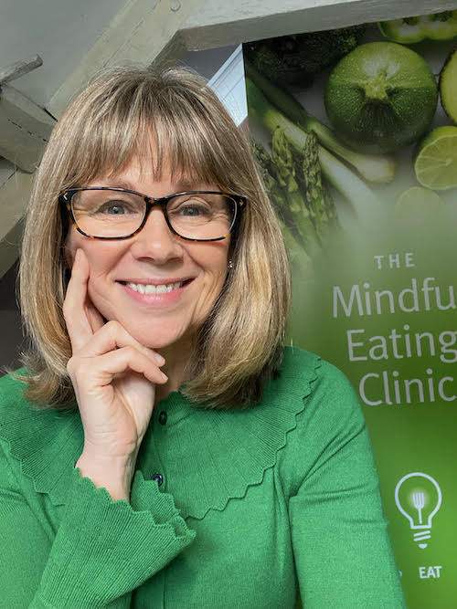 Lori Weber has her right hand to her face and a lovely smile on her face. She has blondish hair to shoulder level and is wearing black rimmed glasses. Behind her left shoulder is her pull up roller banner which is vibrant green and you can make, even though the last letters are missing, that in white is written the mindful eating clinic. She is wearing a bright green top.
