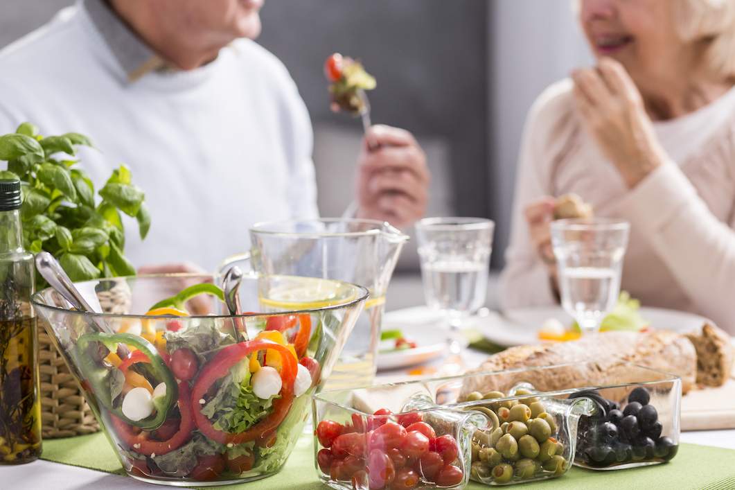 An older couple are sat in the background of the photo they are bot wearing white and eating. In the foreground are glasses of water a bowl of peppers and two other separate glass square bowls one has tomatoes in and the other green olives. They are clearly loving the meal and each others company.