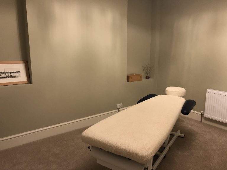 An image of a treatment room that looks very relaxing. It has green walls and wire sculpture of some flowers on a small piece of drift wood and a Bach flower essence box both are on a recessed area. To the right is a treatment couch with a beige cover and blue arm extensions.