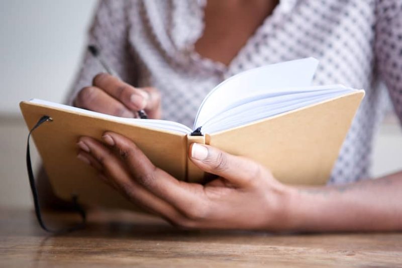 This is a photo of an Indian lady sitting at a table with a cream journal in her hand. She is writing in the book but you can only see her hand holding it. She is wearing a white shirt aith black spots on it.