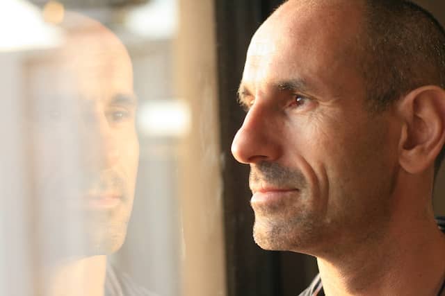 Close up photo of a middle aged man looking out of a window. You can see the reflection of his face in the window and sunlight reflecting on his face. To represent self reflection about possible benefits of things staying the same