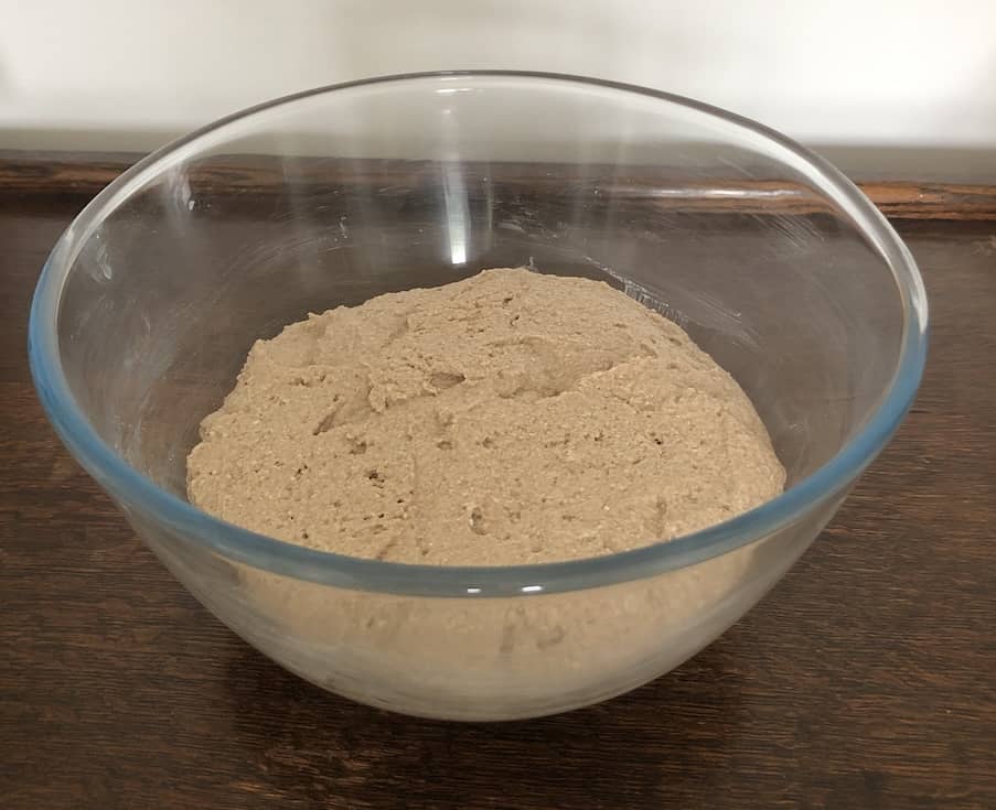 Photo of the mised dough for gluten free teff rolls. It's all mixed in a large glass bowl on a wooden surface ready to be covered to rise.
