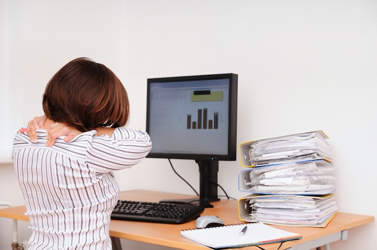 A lady with a pile of paperwork on her desk she is sat facing away looking at her computer monitor that has a graph on it. She is holding her neck to show she is in pain.