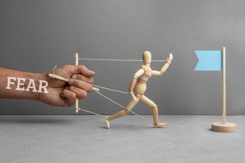a wooden art manequin being held back by puppet strings the person holding it back has the word fear superimposed over the top