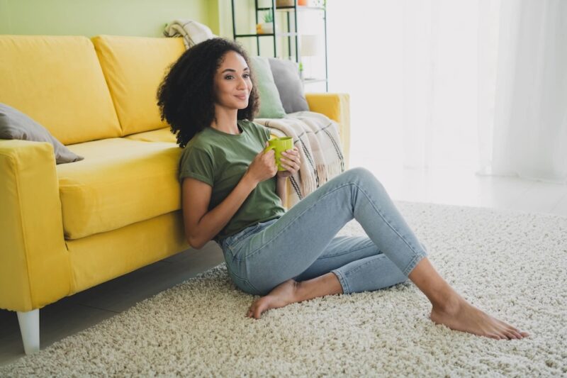 A lady with light brown skin and black curly hair is reclined against a yellow sofa holding a mug, she looks reflective as though she is thinking about something.