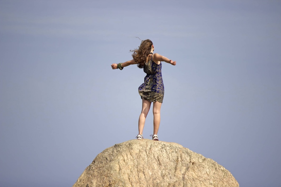 Blue sky. There is a huge rock that a girl is stood on top of with her arms outstretched she is turned away but looks like she's saying yay I'm on top of the world, this is my life, my destiny, my choices. This relates to a blog describing how having a sense of control over your own life is a great immune booster.