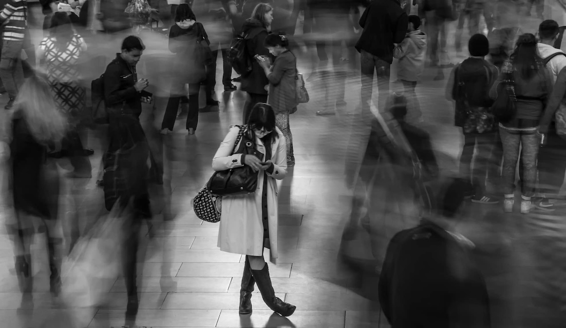 This is black and white photo there is a lady with two bags on her shoulder looking down at her phone. All around her is a busy scene of people rushing around or being stood in queues the photo has movement to it so the detail isn't clear. This is representing how busy we can get in life which has a detrimental impact on our immune systems.